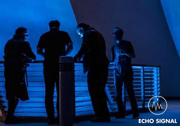 Phaedrus, Adam, John, and Charles (Echo Signal in 2016) photographed from behind, leaning on a railing in heavy blue light.