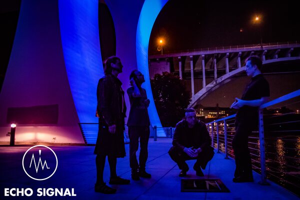 Phaedrus, Charles, John, and Adam (Echo Signal in 2016) under a bridge span at night, lit in magenta and blue, the lit arch of a second bridge behind them.